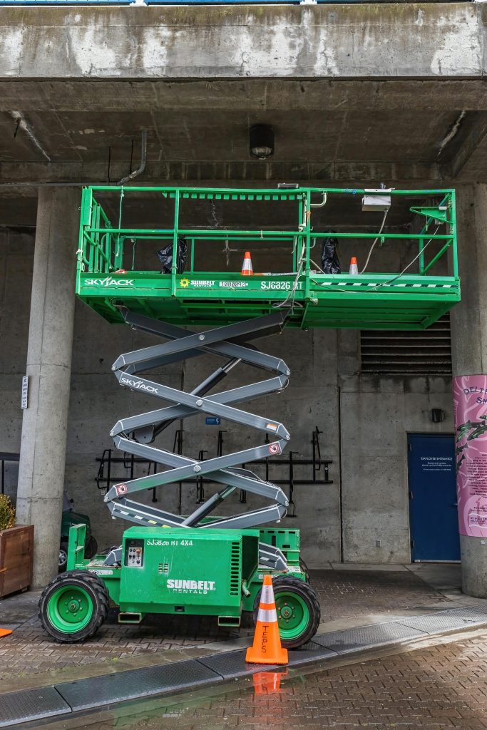 Green scissor lift parked outside an urban building with safety cones around.